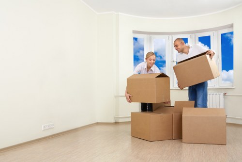 A man and woman are inside an unfurnished, bright room with a large bay window showing a partly cloudy sky outside. The man, dressed in casual light clothing, is in the process of lifting a medium-sized cardboard box, while the woman, also casually dressed, holds a similar box and appears to be preparing to place it on the floor. Several other cardboard boxes of various sizes are positioned on the light wooden floor, some stacked or placed nearby. The room has plain white walls, an electrical outlet visible near the floor, and no furniture or decorations, indicating a space prepared for moving or packing, consistent with house removals services in the Hackney area. The natural daylight from the window illuminates the scene, emphasizing the clean and open environment suitable for a residential relocation process, supported by local removal specialists like Hackney Man with Van.